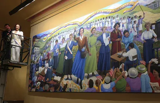 Artist Phyllis Garibay-Coon, in white, stands in front of a maintenance worker on a lift following the unveiling of her mural in the Statehouse honoring women who campaign for decades for voting rights, Wednesday, Jan. 29, 2025, in Topeka, Kan. (AP Photo/John Hanna)