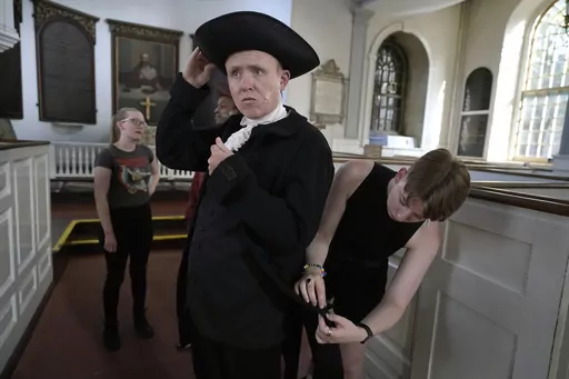 Actor Brooks Reeves, in the role of Mather Byles, center, receives assistance with his costume from co-producer Jess Meyer, right, before the start of a dress rehearsal of the play "Revolution's Edge" Monday, June 12, 2023, at Old North Church, in Boston's historic North End neighborhood. Old North Church, which played a pivotal role in the nation's fight for independence, and is one of Boston's most popular tourist attractions, will host a theater performance for the first time in the church's 
