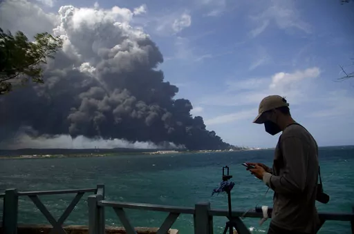 A man types on his cellphone near a huge plume of smoke rising from the Matanzas Supertanker Base as firefighters work to quell the blaze which began during a thunderstorm in Matanzas, Cuba, Monday, Aug. 8, 2022. Cuban authorities say lightning struck a crude oil storage tank at the base, sparking a fire that sparked four explosions. (AP Photo/Ismael Francisco)