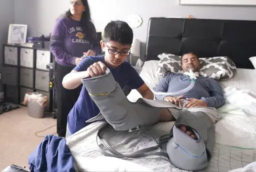 Ronan Kotiya, 11, removes a compression leg sleeve from his father Rupesh Kotiya as his mother Siobhan Pandya looks at their home in Plano, Texas, Sunday, April 10, 2022. Ronan helps care for his father who suffers from ALS and is dependent on a ventilator and around the clock care. Millions of Americans with serious health problems depend on children ages 18 and younger to provide some or all of their care at home. An exact number is hard to pin down, but researchers think millions of children 