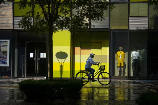 A woman wearing a face mask rides a bicycle past stores closed for COVID-19 control at a shopping mall in Beijing, Tuesday, May 17, 2022. Neither inflation nor the war in Ukraine are threatening to take a bite out of the luxury fashion market, according to a study published Tuesday, June 21, 2022. (AP Photo/Mark Schiefelbein, File)