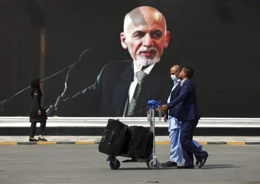 Passengers walk to the departures terminal of Hamid Karzai International Airport in Kabul, Afghanistan, on Saturday, Aug. 14, 2021, past a mural of President Ashraf Ghani, as the Taliban offensive encircled the capital. On the eve of the anniversary of the Taliban takeover of Kabul, Afghanistan's former president on Sunday, Aug. 14, 2022, defended what he said was a split-second decision to flee, saying he wanted to avoid the humiliation of surrender to the insurgents. (AP Photo/Rahmat Gul, File