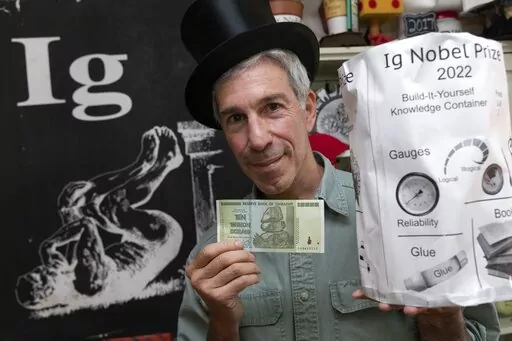 Master of Ceremonies Marc Abrahams poses with the 2022 Ig Nobel prize, Friday, Sept. 9, 2022, at his office in Cambridge, Mass. The prize was emailed in advance to the winners with instructions for self-assembly. The annual prize ceremony on Thursday, Sept. 15, 2022, was for the third year in a row a prerecorded affair because of the lingering effects of the coronavirus pandemic. Abrahams also holds a Zimbabwean $10 trillion bill that is part of the prize presented to the winners. (AP Photo/Mich