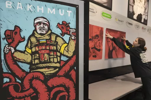 Veronika Yadukha, of Kyiv, Ukraine, a student at Dartmouth College, right, installs an exhibit by Ukrainian artists near a poster called "Bakhmut," by Mykhailo Skop, left, before the opening of an art exhibit called "Our Fire is Stronger Than Your Bombs" at Saint Anselm College, in Manchester, N.H., Monday, May 1, 2023. The art exhibit, marking the one-year anniversary of the Russian invasion of Ukraine, features new work by Ukrainian illustrators, some of whom have been living without electrici